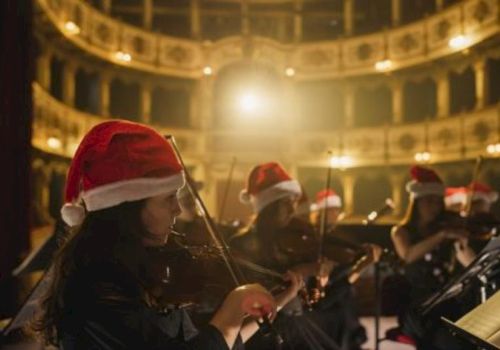 A group of violinists wearing Santa hats perform on a warmly lit stage, ready to play as festive lights glow above the ornate theater.