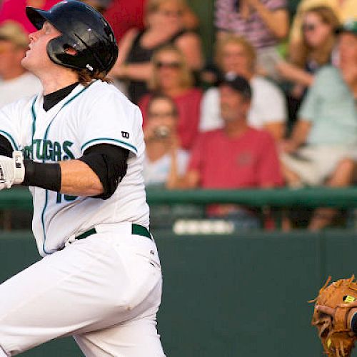 A baseball player swings a bat during a game, watched by a catcher and umpire, with a crowd of spectators in the background.