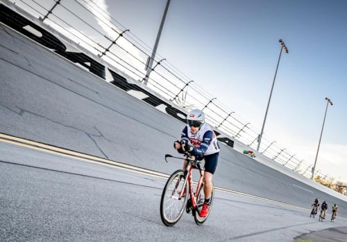 A cyclist in a road-race rides past a concrete wall under overhead power lines while other riders follow behind.