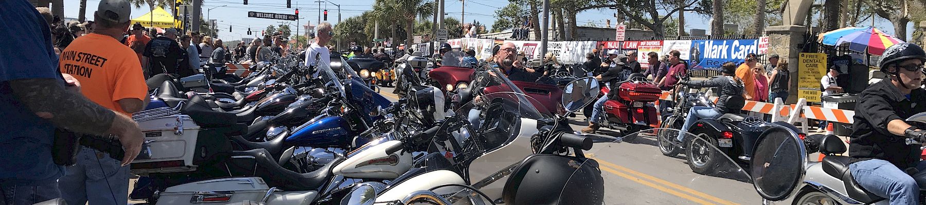 A crowded street lined with motorcycles, people, palm trees, and a banner overhead, indicating a motorcycle event or gathering.