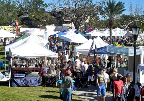 A lively outdoor market with white tents, crowds strolling between stalls, palm trees, and booths selling crafts and foods.