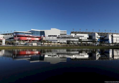 A stadium race track building labeled &ldquo;Daytona International Speedway,&rdquo; reflected in a calm body of water under a clear blue sky.