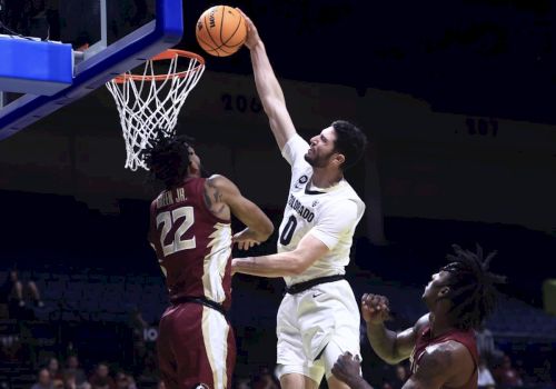 A basketball player in white leaps for a shot as a maroon defender jumps; the ball is near the rim during a game.
