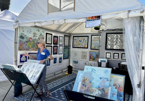 A woman wearing a blue shirt stands in a white tent filled with colorful framed art and quilts displayed on walls and stands.
