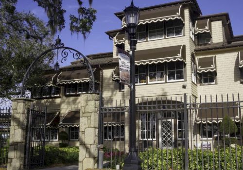 A stately, cream-colored mansion with wrought-iron gates and striped awnings, set behind a stone entrance and a tall street lamp.