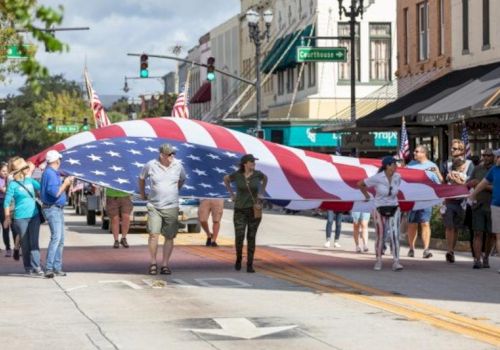 A group of people are carrying a large American flag during a street parade in an urban area with shops in the background.