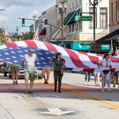 A group of people are carrying a large American flag during a street parade in an urban area with shops in the background.