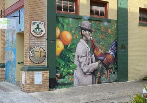 A mural on a building wall depicts a man in a hat holding a rooster, with oranges and leaves in the background.