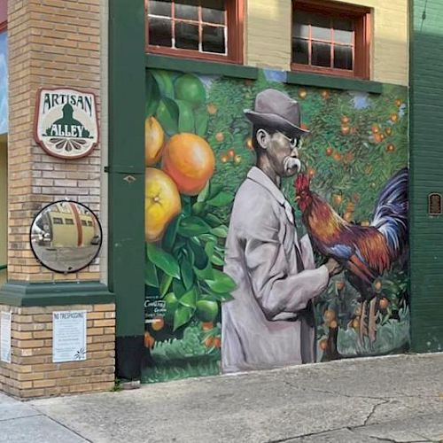 A mural on a building wall depicts a man in a hat holding a rooster, with oranges and leaves in the background.