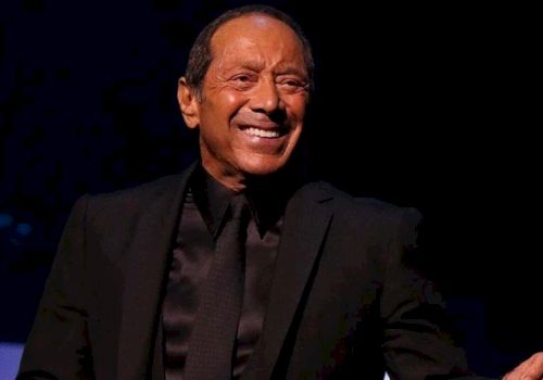 An older man in a dark suit and tie is smiling on stage against a dark background.