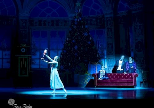 A ballet performance shows a dancer holding a Nutcracker, with a large Christmas tree in the background and two people seated on a couch.