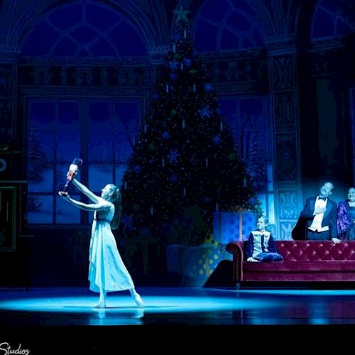 A ballet performance shows a dancer holding a Nutcracker, with a large Christmas tree in the background and two people seated on a couch.