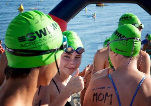 A group of swimmers with green caps, featuring "SWIM ACROSS AMERICA," gather near the water, with one giving a thumbs up.