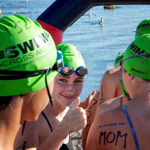 A group of swimmers with green caps, featuring "SWIM ACROSS AMERICA," gather near the water, with one giving a thumbs up.