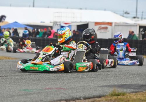 People in go-karts racing on a track, wearing colorful helmets and suits with spectators and tents in the background.