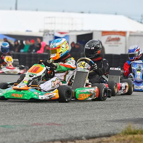 People in go-karts racing on a track, wearing colorful helmets and suits with spectators and tents in the background.