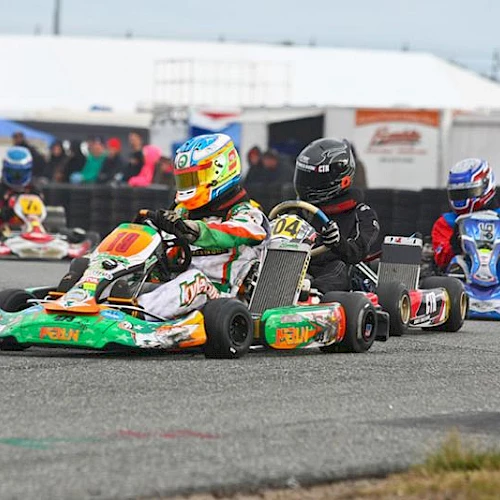People in go-karts racing on a track, wearing colorful helmets and suits with spectators and tents in the background.
