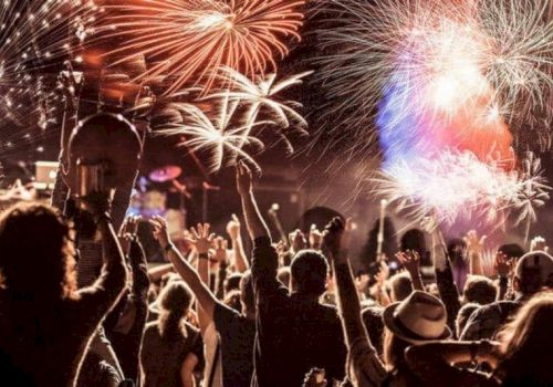 A crowd celebrates under a sky illuminated by colorful fireworks, with people raising their hands, creating a festive atmosphere.