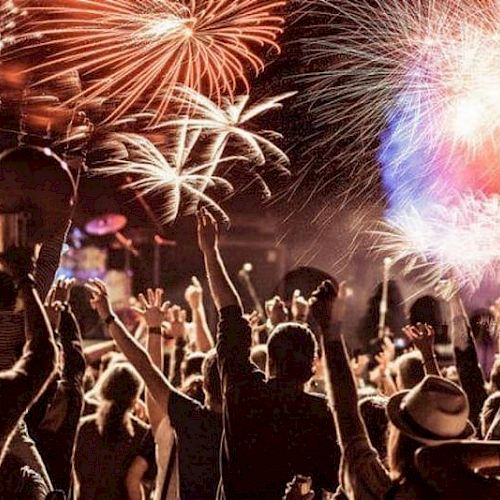 A crowd celebrates under a sky illuminated by colorful fireworks, with people raising their hands, creating a festive atmosphere.