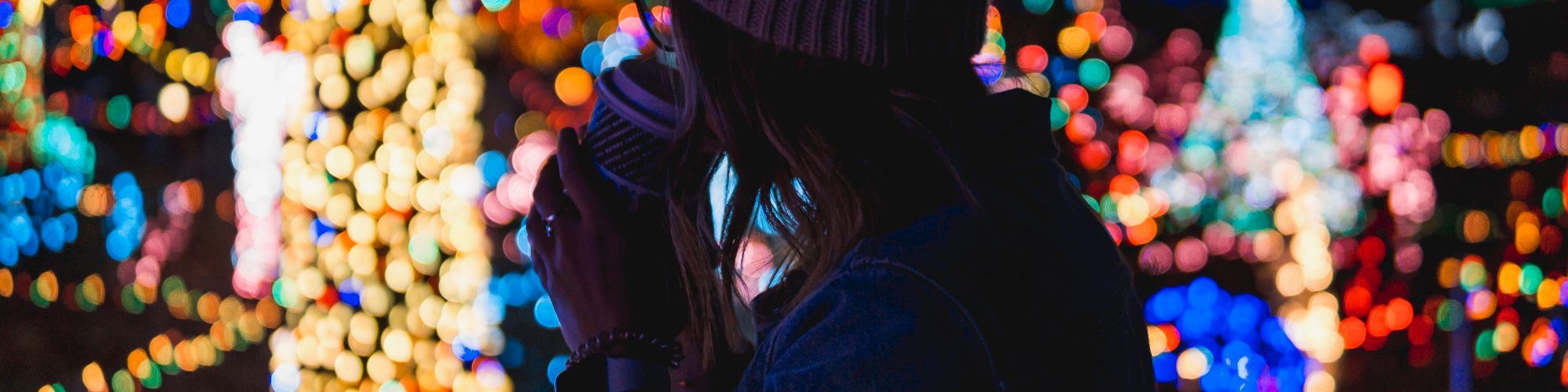 A person in a beanie holds a cup, gazing at colorful, blurred lights in a festive, nighttime setting.