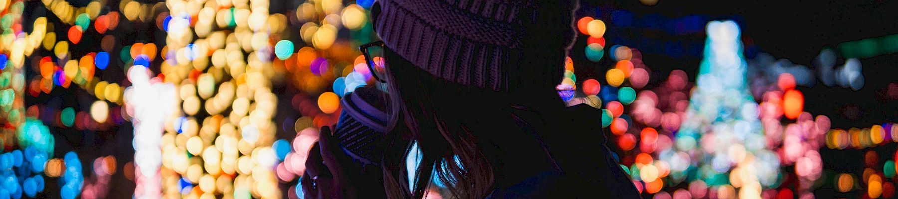 A person in a winter hat enjoys a hot drink while colorful holiday lights glow in the background.