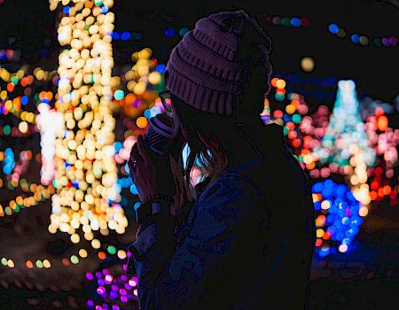A person in a winter hat enjoys a hot drink while colorful holiday lights glow in the background.