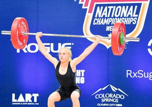 A female weightlifter is lifting a barbell overhead in a competition, with a USA Weightlifting backdrop behind her.