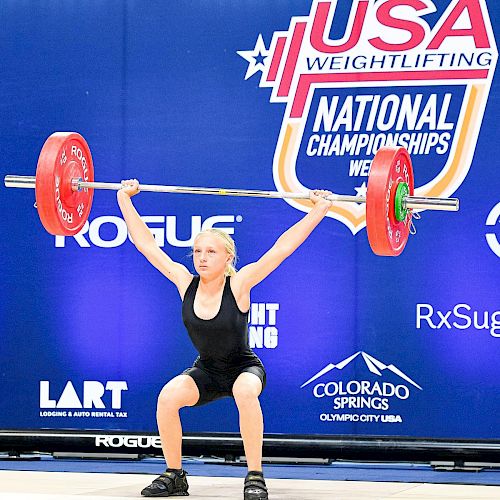 A female weightlifter is lifting a barbell overhead in a competition, with a USA Weightlifting backdrop behind her.