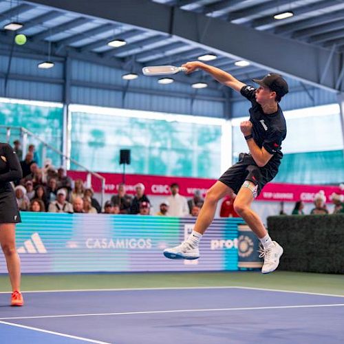 A tennis player in mid-air serving on a blue court, with a referee and crowd watching in a large indoor arena.