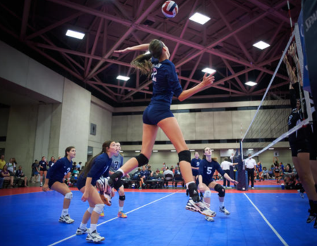 Volleyball action mid-air: a player spikes over the net while teammates and opponents watch on a bright blue court inside a gym.