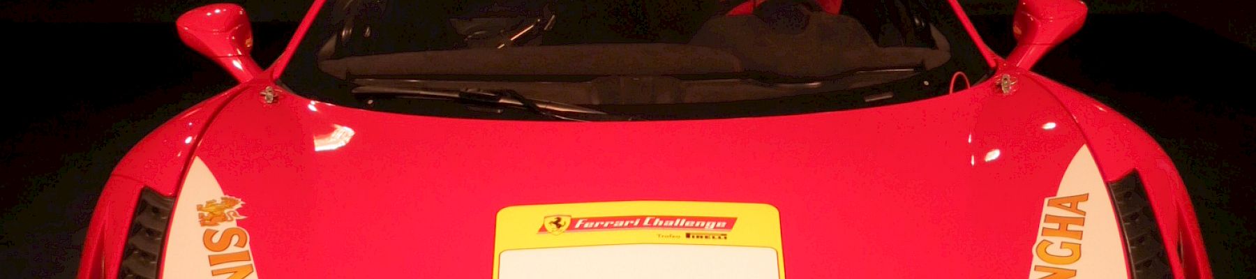 A red Ferrari race car with Pirelli logo, sponsor decals, yellow license area, and showroom lighting in the background.