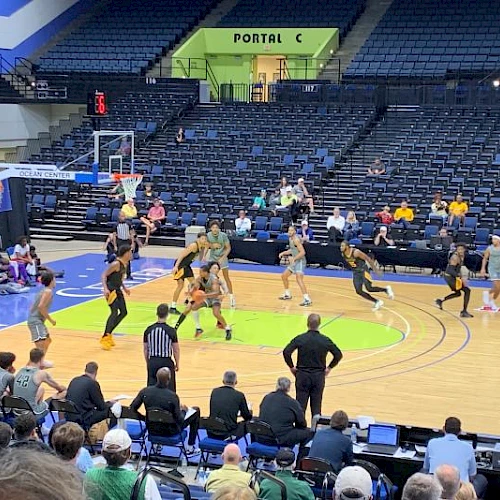 A group of players in a basketball game on a court, with spectators in the stands and a scorer&rsquo;s table nearby, action-packed on the court.