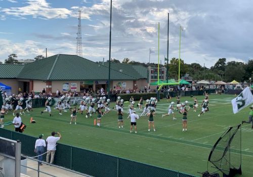 Cheerleaders perform on a football field during a game or event, with spectators, a building, and tall goalposts in the background, sunny skies.