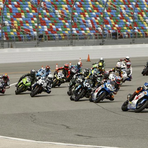 A line of motorcycle racers leaning into a curve on a track, colorful stands in the background, cones near the pit wall, racing action intensifies.