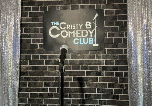 A mic stands on a stage with a sign reading &ldquo;The Cristy B Comedy Club&rdquo; against a brick wall, framed by sparkly curtains.
