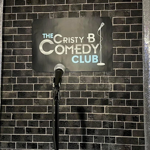 A mic stands on a stage with a sign reading &ldquo;The Cristy B Comedy Club&rdquo; against a brick wall, framed by sparkly curtains.