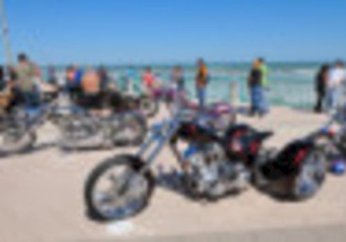 A beach scene with a row of motorcycles parked on sand while people gather near the shoreline, on a sunny day.