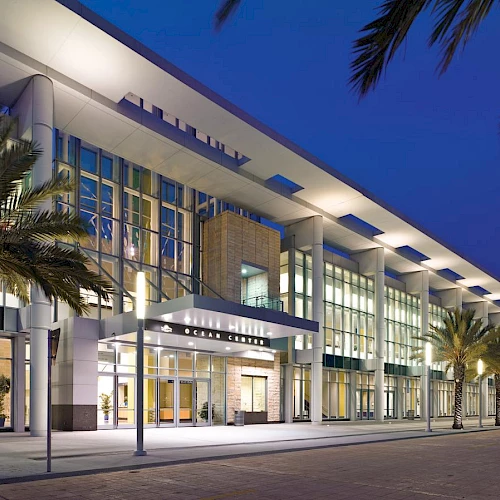 A modern glass-front building at dusk with palm trees lining a well-lit walkway outside, suggesting a shopping or civic complex.