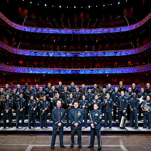 A large formal orchestra sits on stage with conductor in front, many musicians in uniform, a cello on the right, inside a grand theater.