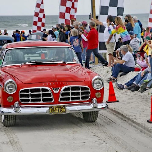 A red vintage car races along a dusty road at a crowd-lined car show, with checkered flags and spectators cheering nearby.
