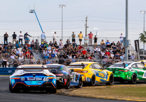 A lineup of race cars on the track with fans in the stands behind them, vehicles ready for a race as spectators watch intently.