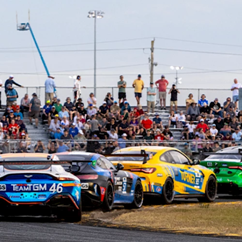 A lineup of race cars on the track with fans in the stands behind them, vehicles ready for a race as spectators watch intently.