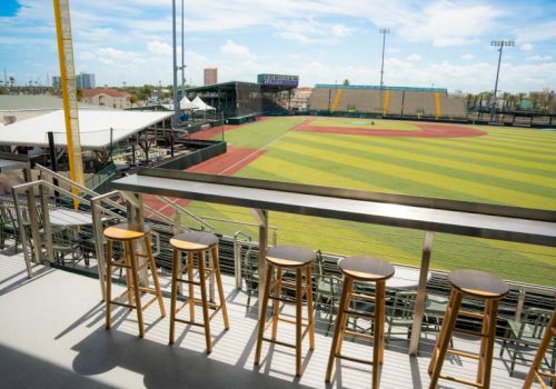 A view of a baseball/softball stadium with a brightly colored turf field and barstools on a balcony overlooking it.