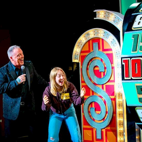 A man and a woman cheerfully pose on a gameshow stage beside a tall, colorful wheel and oversized numbers, enjoying a fun prize moment.