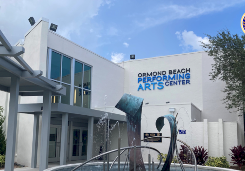 A view of the Ormond Beach Performing Arts Center exterior with a metal water fountain in the front and trees under a blue sky.