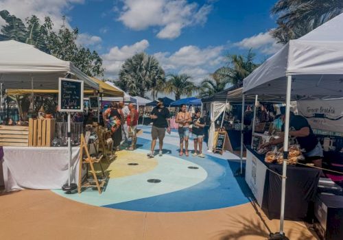 A sunny outdoor market with white tents, vendors selling goods, and people strolling along a colorful painted courtyard.