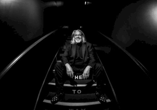 A black-and-white photo shows an older man seated in a dark spotlight on steps, with &ldquo;THE&rdquo; and &ldquo;TO&rdquo; text beneath him.