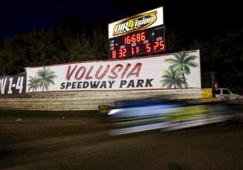 A slow-motion race car blurs by along a dirt track with a bright VOLUSIA SPEEDWAY PARK sign and scoreboard in the background at night.