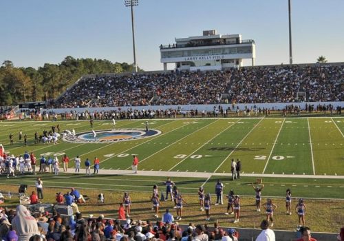 A crowded outdoor football stadium with fans in the stands, a long field, players on the sideline, and a large press box overlooking the end zones.