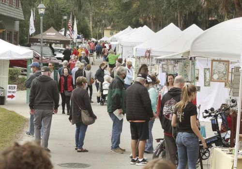 Shoppers browse a row of white tents at an outdoor market or fair, people chatting and looking at goods along a tree-lined path.